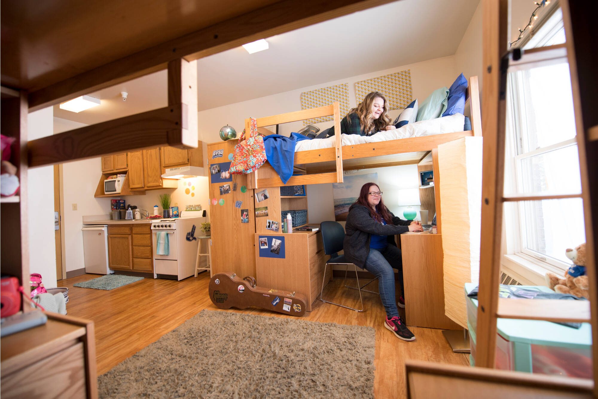 One bedroom apartment style room with a lofted bed and a desk below. One person studies at the desk, while another relaxes above. Warm lighting and a tidy kitchen area are in the background.